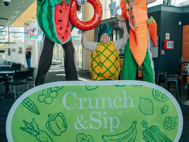Image of Three people dressed in large fruit and vegetable costumes, including a watermelon, carrot, and pineapple, pose and wave behind a green Crunch & Sip display sign in a brightly lit indoor space.