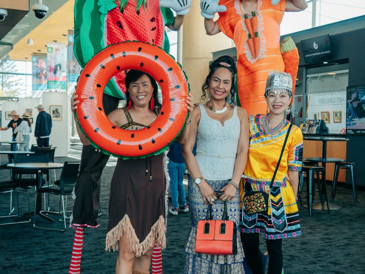 Image of Four people pose indoors whilst two others on stilts wear whimsical fruit and vegetable costumes (watermelon and carrot). The four in front wear a fringed brown dress, a blue lace dress, and a colourful embroidered outfit.