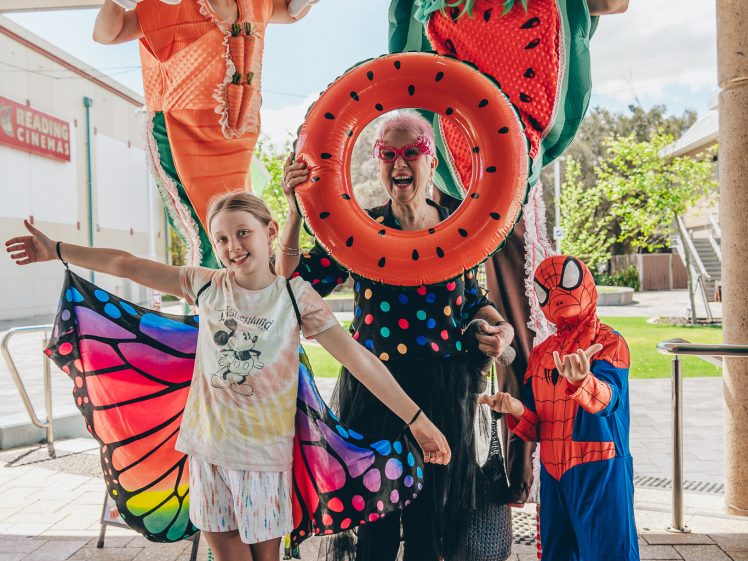 Image of A group of people in colourful costumes pose outdoors. Two adults on stilts wear fruit-themed outfits, one child is dressed as Spider-Man, another as a butterfly, and an older woman stands in the centre wearing a tutu and pink wig.