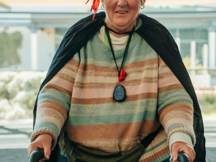 Image of An older woman wearing a pirate hat, striped jumper, and black cloak smiles whilst using a walking frame. She has a green bum bag and a stopwatch around her neck, standing indoors with natural light behind her.