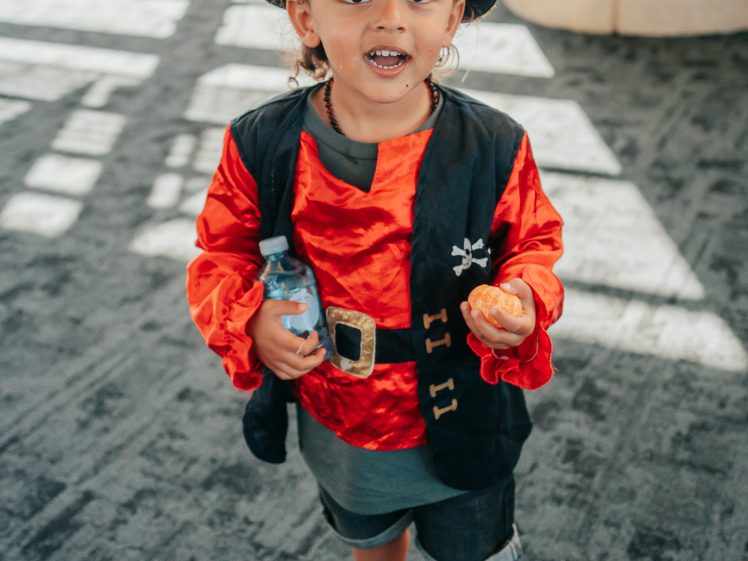 Image of A young child dressed in a pirate costume with a hat and red shirt holds a water bottle and a small snack, standing on a carpeted floor indoors and smiling at the camera.