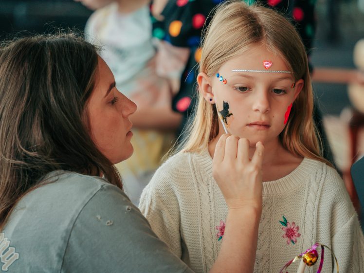 Image of A woman paints a black cat on a young girl's cheek. The girl, wearing a white jumper and decorated with stickers and face paint, sits still and looks ahead while the woman carefully paints her face.