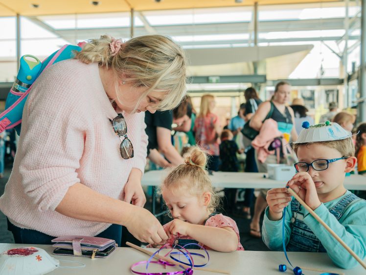 Image of A woman helps two young children with a craft activity at a busy indoor event. The boy wears glasses and a decorated paper plate hat, whilst the girl focuses on threading beads. Other people and children are visible in the background.
