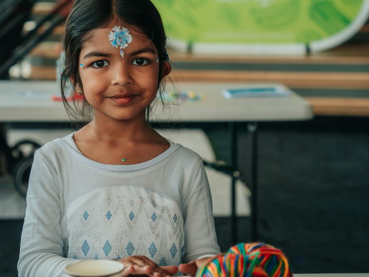 Image of A young girl with a jewelled blue bindi and face stickers sits at a craft table with a colourful yarn ball, wooden sticks, and paper shapes. She smiles softly, with stairs and a Crunch & Sip sign in the background.