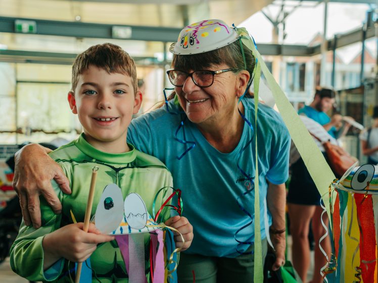 Image of A smiling boy in a green costume holds a colourful craft with ribbons, standing next to an older woman in glasses and a blue shirt, who is also smiling and has her arm round him. They are indoors at a festive event.