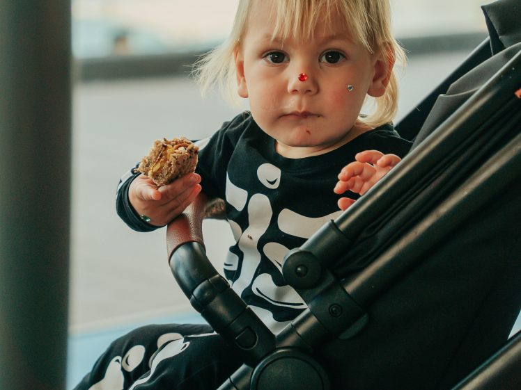 Image of A toddler wearing a black outfit with a white skeleton pattern sits in a pushchair, holding a piece of food. The child has blonde hair and a few small stickers on their face. The background is softly blurred.
