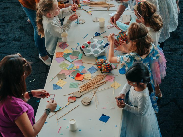 Image of Children and adults gathered around a table covered with craft supplies, such as coloured paper, sticks, and paint, working together on art projects in a lively, creative setting.
