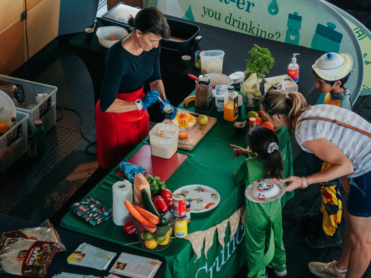 Image of A woman in a red apron prepares food at a table while two children and a woman watch. The table is covered with ingredients, utensils, and vegetables; educational materials are displayed around them.