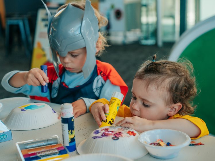 Image of Two young children sit at a table making crafts with paper plates and glue sticks. One child wears a Thor helmet fancy dress, and both are focused on decorating their paper plates with colourful art supplies.