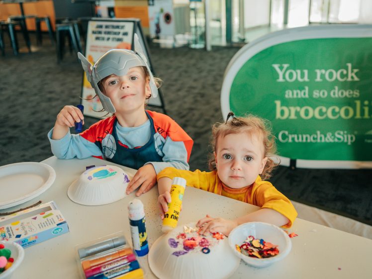 Image of Two young children sit at a table doing crafts with paper plates, glue, and colourful decorations. One wears a Thor costume and helmet. Behind them is a green sign that reads, “You rock and so does broccoli! Crunch&Sip.”.