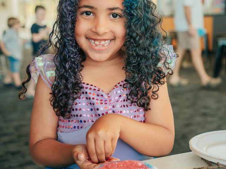Image of A young girl with curly hair, partly dyed blue, smiles whilst decorating a bowl with colourful glitter at a table indoors. She is wearing a purple sequinned dress. Other people are blurred in the background.