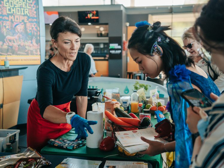 Image of A woman wearing gloves explains food items on a table to a group of people. The table is filled with vegetables, groceries, and kitchen supplies. A colourful poster is visible in the background.
