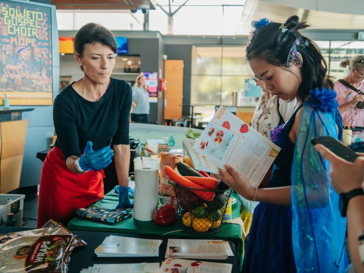 Image of A woman wearing gloves stands behind a table with fruit and vegetables, talking to a young woman holding a leaflet, in a brightly lit indoor setting with a colourful poster in the background.