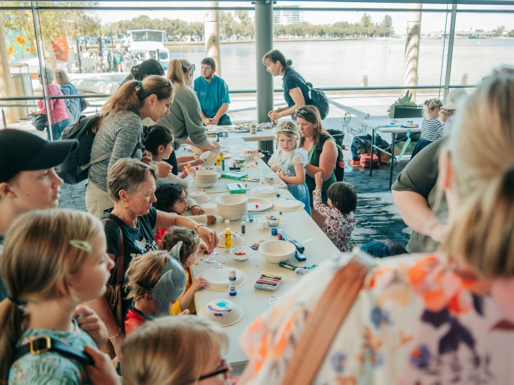 Image of A group of children and adults gather around a long table covered with art materials, engaging in a craft activity in a bright room with large windows overlooking water.