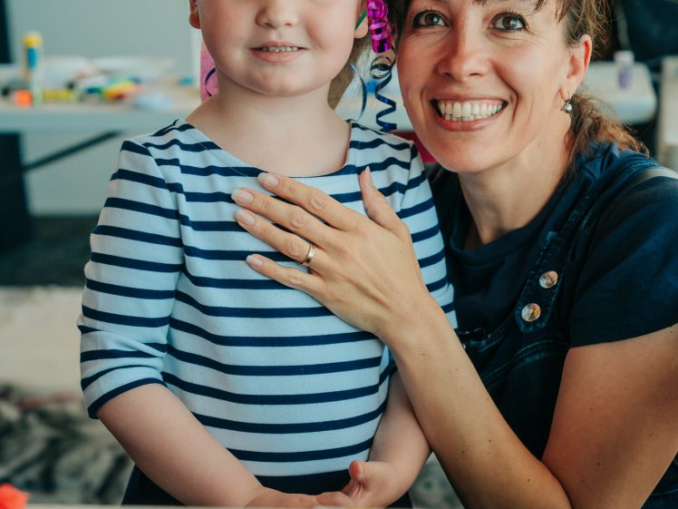 Image of A smiling woman hugs a young girl with decorated face and hair, wearing a striped jumper, at a colourful craft table. Both look happy, surrounded by art supplies in a bright, lively room.