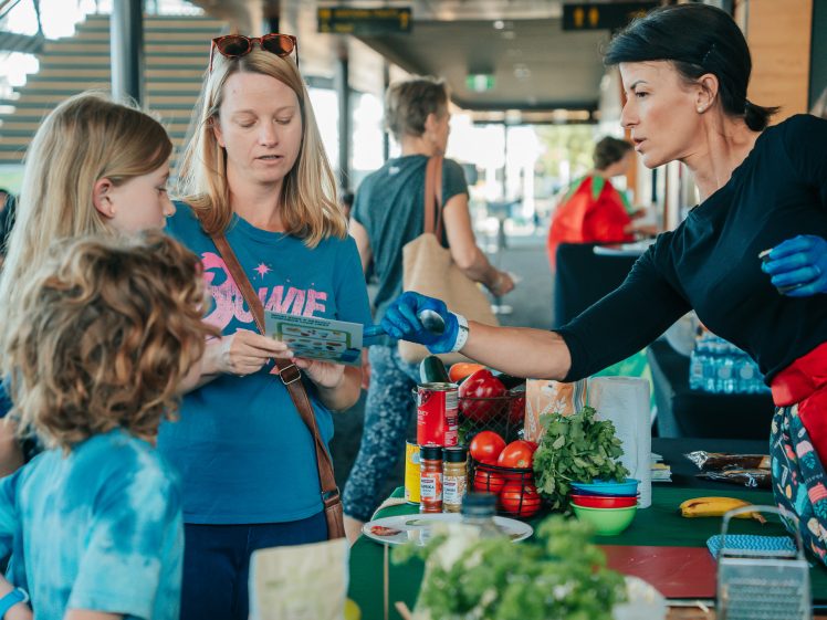 Image of A woman wearing gloves explains something to a woman and two children at a table with fresh vegetables, herbs, and cooking ingredients at an indoor market or event. Other people browse in the background.