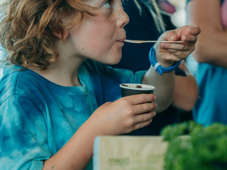Image of A young child with curly hair and a blue shirt tastes food from a small cup, holding up a spoon. Other children are nearby, and there are herbs and a bag on the table in front of them.