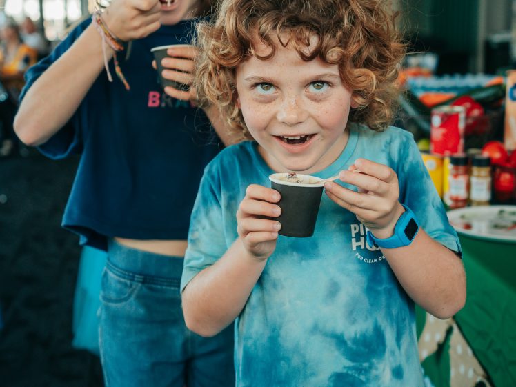 Image of Two children are smiling and eating pudding from cups indoors. The child in front has curly hair and wears a blue tie-dye shirt. The child behind has straight hair and wears a navy shirt. A table with snacks is visible in the background.