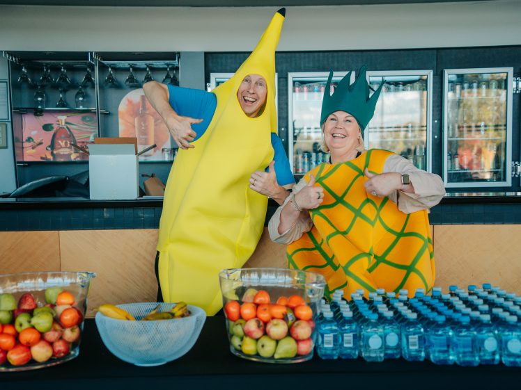 Image of Two people dressed in banana and pineapple costumes smile and give thumbs up behind a counter with bowls of fruit and bottles of water in front of them. Fridges and glasses are visible in the background.