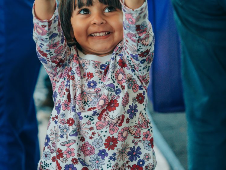 Image of A young child with dark hair, wearing a long-sleeved floral shirt and navy trousers, smiles widely and points both index fingers upward. Other people are visible in the blurred background.