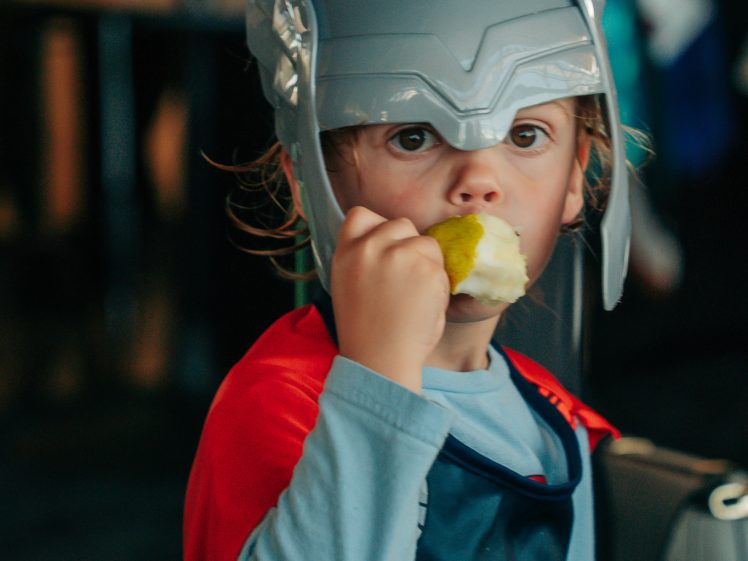 Image of A young child dressed in a superhero costume and helmet, resembling Thor, eats a partially peeled apple whilst looking at the camera. The background is blurred with indoor seating visible.
