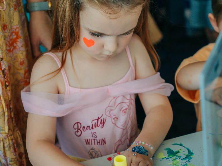 Image of A young girl with a flower hairband and heart face paint draws on colourful paper at a table, wearing a pink dress with “Beauty is Within” text and beaded bracelets. Other children are nearby.