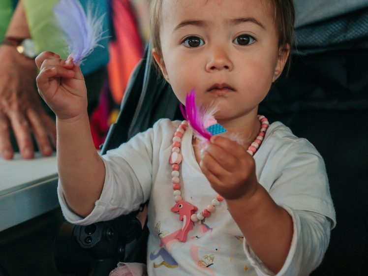 Image of A young child wearing a pink tutu and beaded necklace sits in a pushchair, holding a purple and a blue feather in each hand, looking directly at the camera.