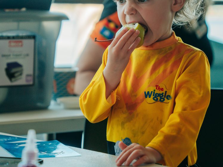 Image of A young child wearing a yellow The Wiggles shirt takes a bite of a green apple whilst sitting at a table with art supplies and colourful paint cups.