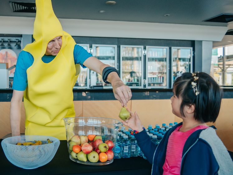 Image of A man dressed in a banana costume hands a pear to a young girl at a table with bowls of fruit and bottles of water indoors.