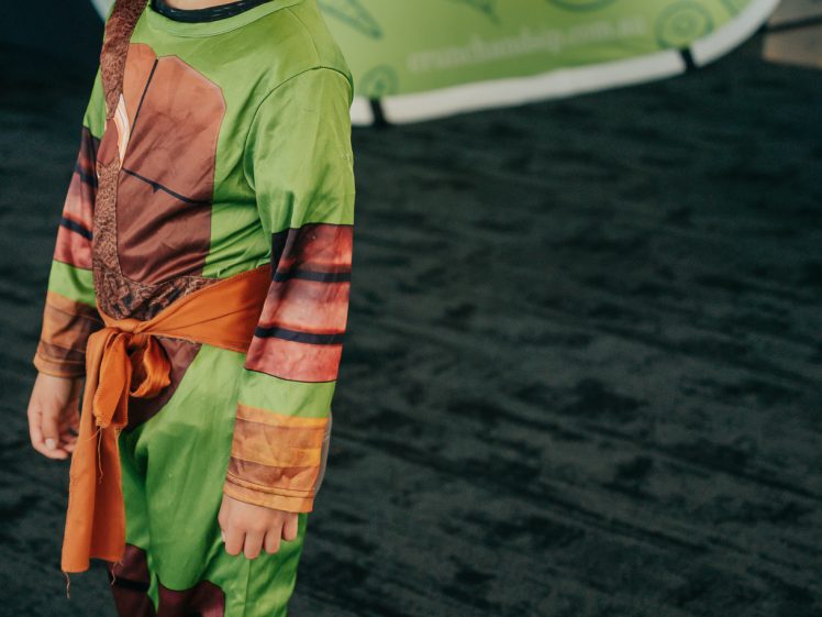 Image of A young child in a colourful superhero costume and painted mask stands on a dark carpet. Behind them is a green Crunch & Sip sign with fruit illustrations.