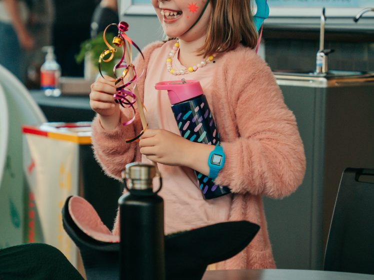 Image of A young girl wearing face paint, a pink cardigan, and a handmade hat smiles whilst holding a colourful ribbon wand and a water bottle at an indoor event. A pirate hat and water bottle are on the table in front of her.