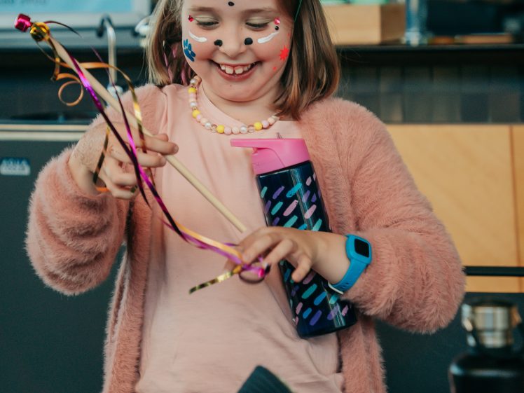 Image of A smiling young girl with face paint and a colander on her head holds colourful ribbons and a water bottle, dressed in a pink cardigan and tutu, enjoying a playful moment indoors.
