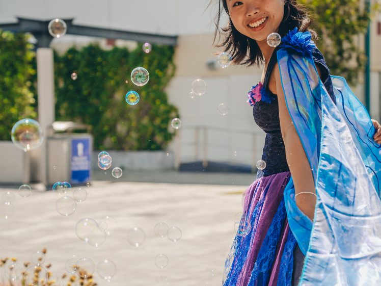 Image of A smiling woman in a blue and purple fairy costume stands outdoors among floating bubbles, with greenery and a building in the background.