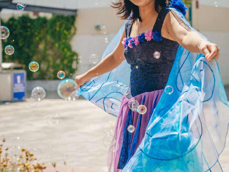 Image of A smiling woman in a blue and purple dress stands outdoors, playfully holding flowing blue fabric as bubbles float around her on a sunny day.