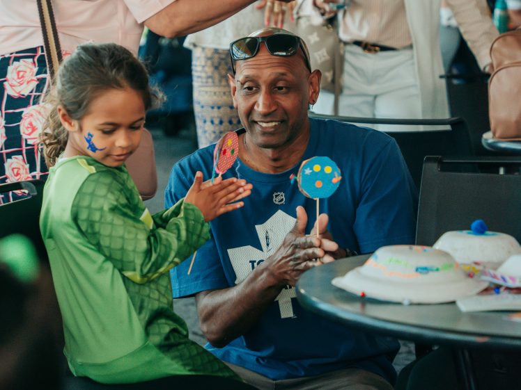 Image of A man and a young girl with face paint smile and clap hands together at a table, holding decorated paper crafts. Other people are gathered round them in the background.