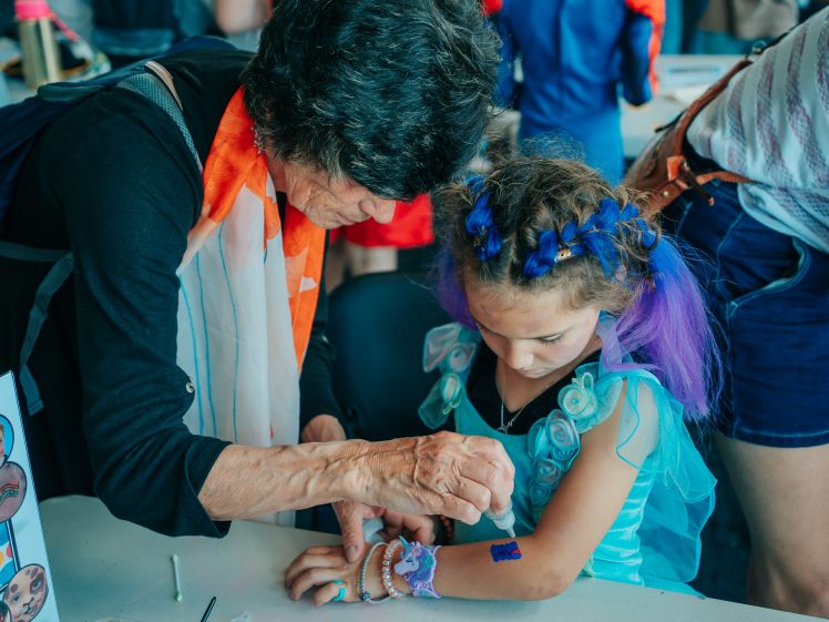 Image of An older woman helps a young girl with blue and purple hair in a fairy costume apply a temporary tattoo to her arm at a table during a festive event.