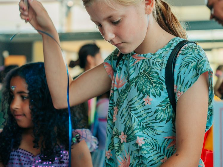 Image of A young girl wearing a glittery, colourful hat and a tropical print dress curls a ribbon at a busy indoor event. Another child in a purple outfit is nearby, and people mingle in the background.