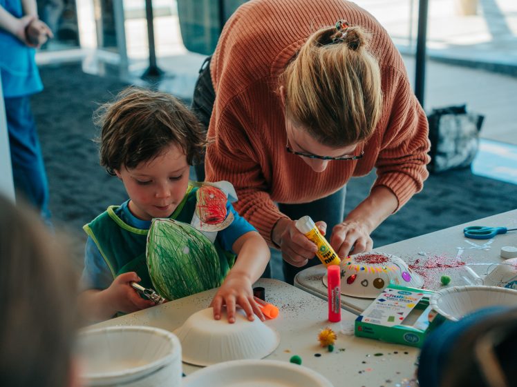 Image of A woman helps a young child decorate paper plates with craft supplies like glue, felt-tip pens, and pom-poms at a table during an arts and crafts activity.