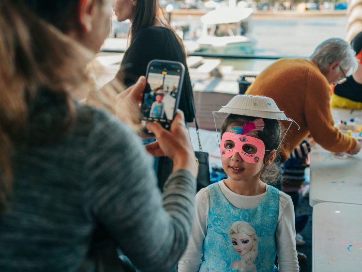 Image of A child in a blue Frozen dress, pink mask, and a paper hat is being photographed by an adult with a mobile phone at a craft table by the water. Other people are also crafting in the background.