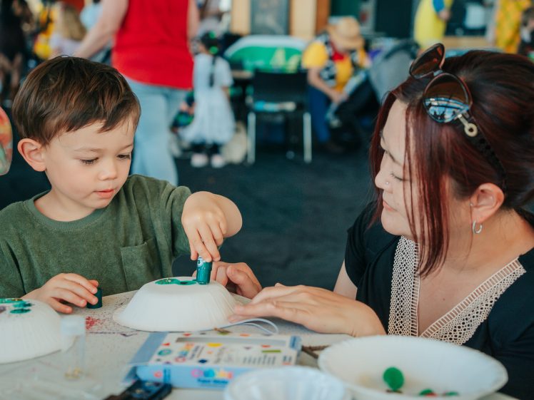 Image of A young boy and a woman sit at a table, painting and decorating white paper bowls together. The boy focuses on his craft whilst the woman watches, both engaged in a creative, hands-on activity.