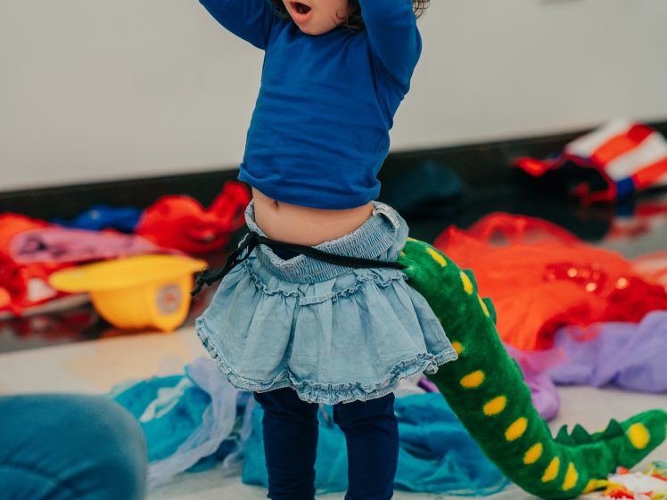 Image of A young child in a blue shirt, denim skirt, and dinosaur tail stands on a floor covered with colourful costumes, holding a yellow hard hat above their head with a surprised expression.