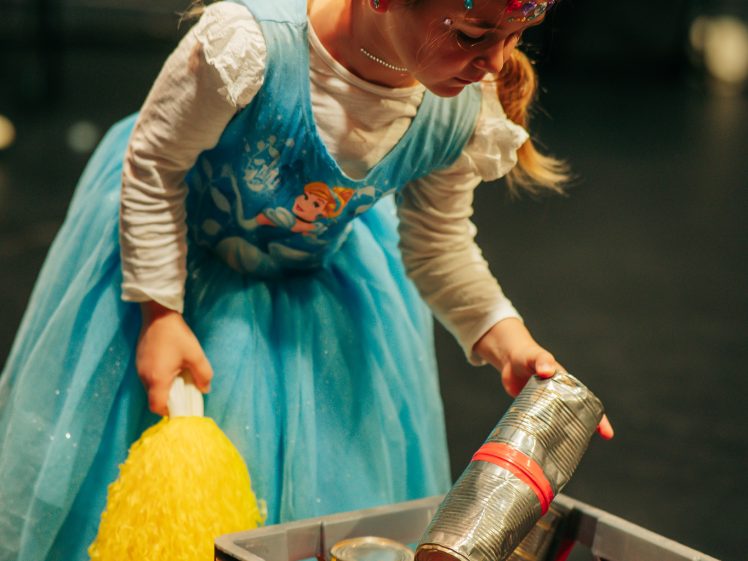 Image of A young girl in a blue princess dress holds a yellow pom-pom and reaches into a crate filled with tin cans. She has face paint and is focused on the activity, with blurred people and chairs in the background.