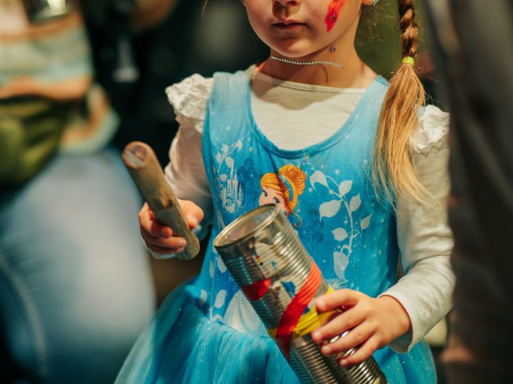 Image of A young girl in a blue princess dress holds a metal tin and a stick, ready to play music. She has colourful heart stickers on her forehead and a red star painted on her cheek, looking focused in a crowded room.