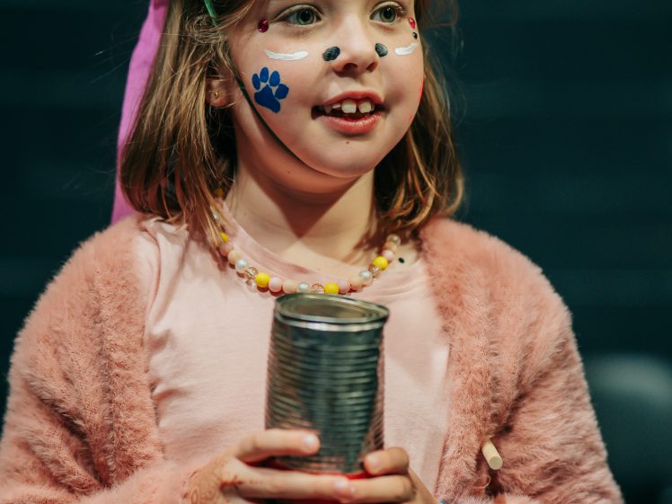 Image of A young child wearing a fluffy pink jacket, beaded necklace, and decorated hat holds a tin can. Her face is painted with colourful designs and stickers, and she has a blue wristwatch on her left wrist.
