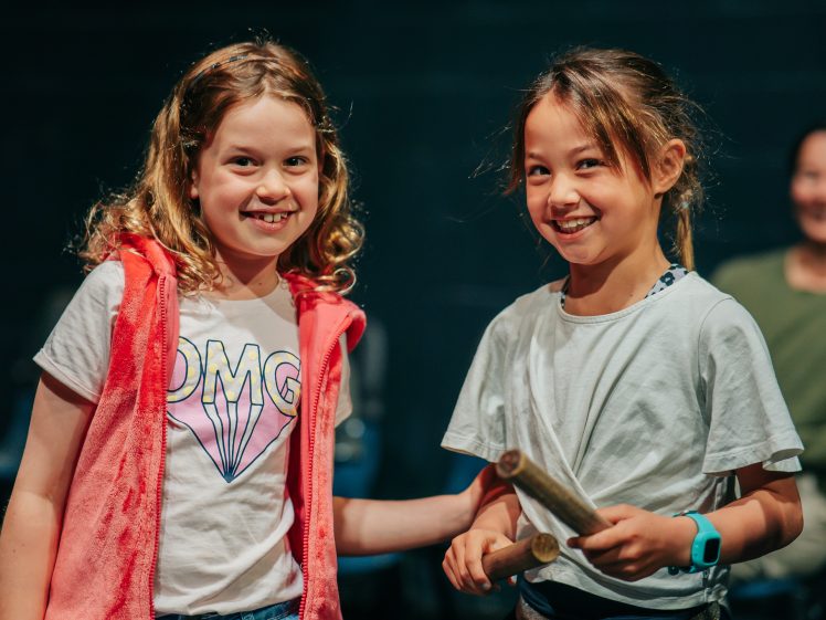 Image of Two young girls smile together. One wears a white OMG shirt with a pink waistcoat, and the other wears a white shirt and holds two wooden sticks. An adult in the background is out of focus.