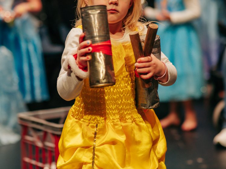 Image of A young girl in a yellow dress holds tin cans and wooden sticks, looking at the camera. Other children in fancy dress stand blurred in the background.