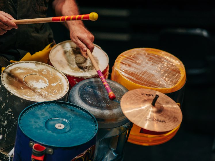 Image of A person plays a drum kit made from colourful plastic buckets, metal containers, and cymbals using two drumsticks, with the focus on the hands and improvised instruments.