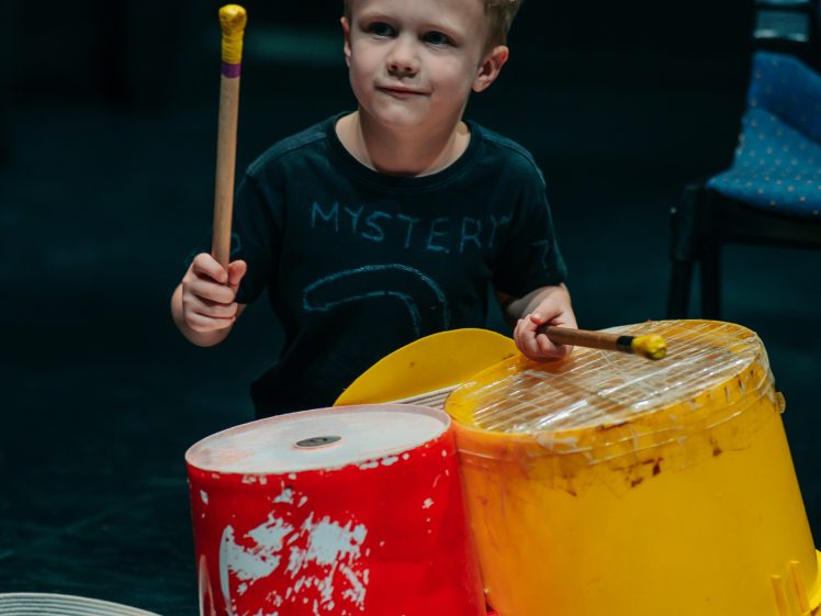 Image of A young boy plays on red and yellow plastic buckets with drumsticks, using them as makeshift drums. He is focused and wearing a dark T-shirt, sitting on a stage or performance area with a chair in the background.
