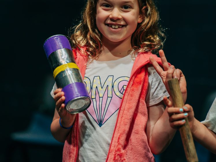 Image of Smiling young girl wearing a pink waistcoat and OMG T-shirt holds a homemade shaker instrument and stands indoors, flashing a peace sign with her other hand.