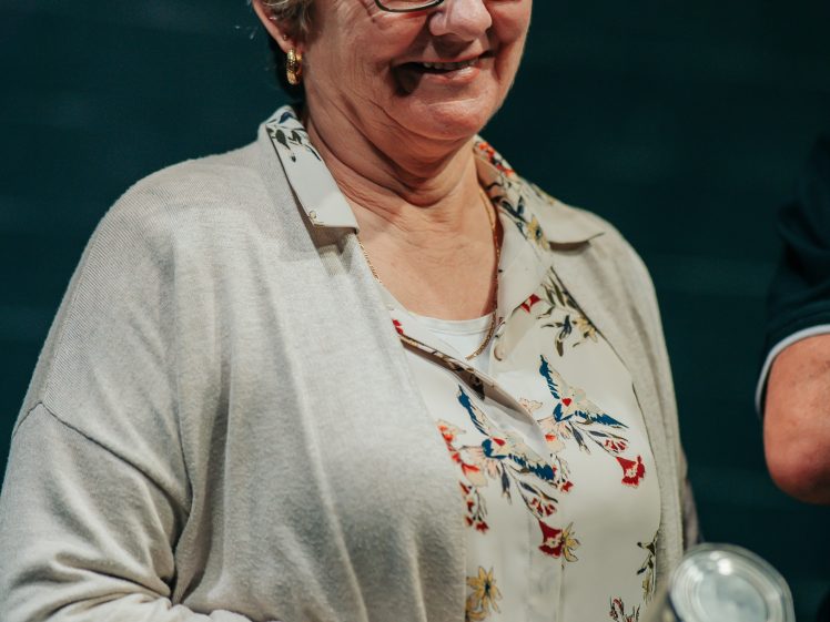 Image of An older woman with short blonde hair and glasses, wearing a light cardigan over a floral blouse, smiles while holding a silver cylindrical object.